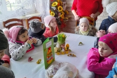 Dzieci siedzą wokół stołu między nimi stoo pani sołtys, na stole leży stroik i cukierki dla dzieci.