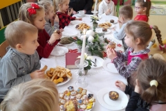 Na zdjęciu widać wigilijny stół przy którym siedzą dzieci i ksiądz. Stół przykryty jest białym obrusem. Na stole widać świece,stroik, sianko, opłatki, gwiazdy betlejemskie i ciasteczka którymi częstują się dzieci.