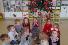 Na zdjęciu widać ustrojona bombkami choinkę. Wokół choinki siedzi grupa dzieci, śpiewa kolędy. Jedna dziewczynka ma na głowie opaskę z Mikołajami. W tle widać kolorowe przedszkolne szafeczki na których umieszczone są zabawki.