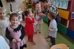 Wiesława Patoń Na zdjęciu widać grupę dzieci składających sobie życzenia świąteczne. Wychowawczyni łamie się opłatkiem z dziewczynka w czerwonej sukience. W tle widać kolorowe prace dzieci na parapecie okien.