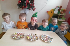 Na zdjęciu przy stoliku siedzi 6 dzieci. Jedna dziewczynka ma na oczach czerwone ozdobne okulary, a na głowie opaskę z choinka. Na stole przed dziećmi na 3 talerzach leżą udekorowane przez dzieci ciasteczka. W tle widać ubrana w kolorowe bombki choinkę i kącik przyrody w postaci języka.