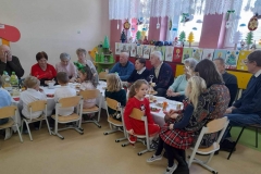 Seniorzy siedzą po jednej stronie stołów, po drugiej dzieci.