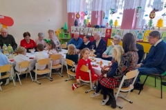 Przy stolikach siedzą Seniorzy  naprzeciwko siedzą dzieci.