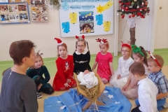 Dzieci siedzą wokół żłóbka z wychowawczynią, w tle kolorowa tablica i choinka.