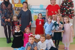 Grupa dzieci stoi wraz z panią sołtys, wychowawca wokół żłóbka. Parę dzieci kuca. Jedna dziewczynka trzyma w reku stroik.