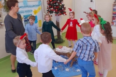 Grupa dzieci tańczy wkoło żłóbka z wychowawczynią, w tle choinka i kolorowa tablica.