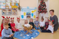 Dzieci siedzą wokół żłóbka, w tle choinka i kolorowa tablica.
