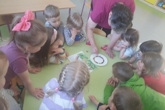 Sala przedszkolna . Grupa dzieci ubrana na kolorowo siedzi przy sześciokątnym zielonym stoliku. Przy stoliku dorosła osoba – wychowawczyni układa na białym talerzyku kolorowe kuleczki na kształt koła. Obok talerzyka rysunek kolorowej tęczy.