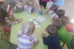 Sala przedszkolna . Grupa dzieci ubrana na kolorowo siedzi przy sześciokątnym zielonym stoliku. Przy stoliku dorosła osoba – wychowawczyni trzyma w ręce plastikową butelkę z wodą nad talerzykiem z kolorowymi kulkami . Dzieci kierują wzrok na talerzyk.