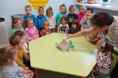 Dzieci zgromadzone przy zielonym sześciokątnym stoliku . Na środku stolika papierowy wulkan , i plastikowe palmy. Osoba dorosła nalewa do otworu wodę .