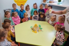 Dzieci zgromadzone przy zielonym sześciokątnym stoliku . Na środku stolika papierowy wulkan , i plastikowe palmy. Z wulkanu zaczyna wypływać żółta ciecz. Na twarzach dzieci zdziwienie i radość.