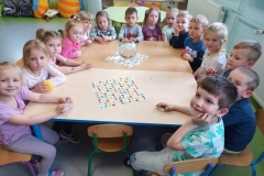 Dzieci zgromadzone przy dwóch jasnych stolikach . Na stoliku szklane naczynie w kształcie kuli wypełnione wodą. Każde dziecko trzyma w dłoni przedmiot np.; papierową łódkę, piłkę, spinacz itp. Za dziećmi widać część sali przedszkolnej z mebelkami , pszczółkami i wózkami dla lalek.