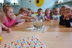 Dieci siedzą wokół stolika że szklaną kulą . Jedno dziecko- dziewczynka wrzuca papierową łódkę do szklanej kuli. Pozostałe dzieci obserwują .