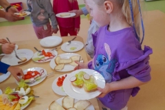 Dzieci stoją wokół stołu, w rękach trzymają talerzyki. Na stole na talerzach leży ułożona wędlina  ser żółty oraz kolorowe warzywa.