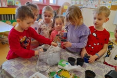 Kilkoro dzieci stoi przy stole na którym leży ziemia, nasiona warzyw i doniczki. Chłopiec w czerwonej bluzce wkłada Ziemię do doniczki.
