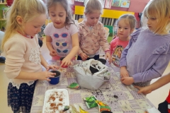 Pięć dziewczynek stoi przy stole na którym leży ziemia  doniczki i torebki z nasionami warzyw. Dwie dziewczynki wkładają Ziemię do doniczki.