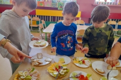 Trzech chłopców stoi przy stole, robią wiosenne kanapki. Na talerzach leżą warzywa, wędlina i serek żółty.