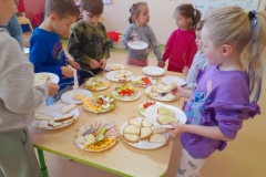 Dziewczynki stoją przy stole, robią wiosenne kanapki. Na talerzach leży chlebek posmarowany masłem, wędlina, ser żółty oraz warzywa.
