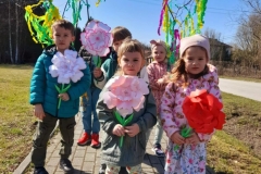 Kilkoro dzieci idzie chodnikiem, dwoje z nich trzyma gaik, reszta wiosenne kwiaty.