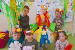 Grupa dzieci, troje z nich siedzi na podłodze trzymają w ręku kwiaty, dwoje w tyle trzyma gaik,  dziewczynka w środku trzyma Panią Wiosnę.