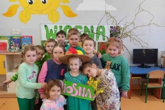 Sala przedszkolna . Dzieci ubrane na zielono stoją w zwartej grupie. Wsród nich dziewczynka ubrana na zielono w słomianym kapeluszu z kwiatami na głowie trzyma w rękach napis WIOSNA z różowych liter na zielonym tle . Dzieci przytulają się do dziewczynki w zielonej sukience. Za dziećmi widać fragment tablicy z napisem WIOSNA , z prawej strony gałązki wierzbowe z zielonymi małymi listkami.