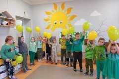 Sala przedszkolna . Dzieci ubrane na zielono stoją w półkolu. Każde dziecko wznosi do góry balonik w kolorze żółtym lub zielonym . W tle za dziećmi widać część tablicy z napisem WIOSNA , fragmenty kolorowych obrazków ,oraz meble przedszkolne.