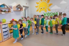 Sala przedszkolna . Dzieci ubrane na zielono stoją w szeregu . Pierwsze dziecko to dziewczynka w kapeluszu i w zielonej sukience trzyma w rękach forsycję. Dzieci poruszają się trzymając między sobą - bez użycia rąk balony na wysokości klatki piersiowej