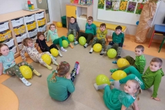 Dzieci siedzą w kole na podłodze trzymają stopami balony zielone lub żółte. Za dziećmi w tle z lewej strony widać szafki na półki indywidualne z prawej strony papierowe drzewo z wierzbowymi gałązkami , a w środku biała tablica z ilustracjami wiosennych kwiatów i półka z bajkami dla dzieci.
