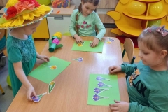Przy prostokątnym stoliku 3 dziewczynki ubrane na zielono ; dwie z nich siedzą , a jedna stoi z lewej strony . Dziewczynki układaja papierowe sylwety wiosennych kwiatów na zielonych kartkach papieru.