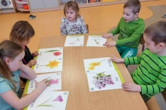 Przy prostokątnym stoliku 3 dziewczynki ubrane na zielono i dwóch chłopców oglądają kolorowe ilustracje z wiosennymi kwiatami. Za dziećmi widać szafki przedszkolne z zabawkami.