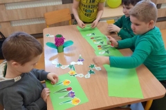 Przy prostokątnym stoliku 4 chłopców ubranych na zielono. Chłopcy układają papierowe sylwety wiosennych kwiatów na zielonych kartkach papieru.