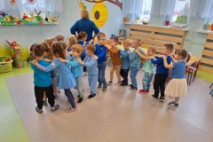 Dzieci w niebieskich ubraniach ustawione wężem za panią. Ręce trzymają na ramionach kolegi. W tle okno, na firanach zawieszone kwiaty.