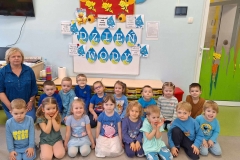 Grupa dzieci ubrana głównie w kolorze niebieskim klęczy na szarej podłodze.Obok nich pani w niebieskiej bluzce. Za nimi na białej tablicy napis na  kropelkach Dzień Wody. Przy napisie hasła o wodzie.