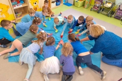 Na podłodze rozłożone błękitne płótno. Wokół niego w kole siedzą dzieci, które pochylają się do środka. W rękach trzymają papierowe, kolorowe łódki. Pod ścianą ustawione pojemniki z klockami i wózki dla lalek.