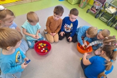 9 dzieci siedzi w półkolu. Przed nimi ustawione plastikowe miski z wodą, w której pływają kolorowe zakrętki. Niektóre dzieci trzymają w rękach klamerki do bielizny. 3 dzieci pochyla się nad miską i próbuje wyłowić klamerkami zakrętki.