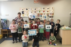 W sali przedszkolnej widoczna jest ustawiona na tle ściany grupa przedszkolaków. W pierwszym rzędzie w środku klęczy chłopiec ubrany w ciemną bluzę i spodnie.       W ręku trzyma transparent z napisem czarnymi drukowanymi literami: Światowy Dzień Wody. Dzieci będące obok niego trzymają w rękach butelki z wodą przewiązane czerwoną kokardą. Dzieci stojące za nimi trzymają plansze - obrazki przedstawiające morze, jezioro i rzekę oraz napis : woda. Troje dzieci w głębi trzyma Transparenty z dużą niebieską kroplą i nieczytelnymi napisami. Kilkoro dzieci ma butelki z wodą. Wśród dzieci po lewej stronie stoi nauczycielka w bluzce w kwiatowy wzór.