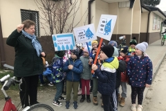 Na tle brązowo - beżowego budynku stoi grupa kilkunastu przedszkolaków. Trzymają w rękach transparenty o treści: Światowy Dzień Wody, Woda źródłem życia, Woda energii doda. Napisy wykonane są czarnymi drukowanymi literami na białym tle. Są tam też duże niebieskie uśmiechnięte krople wody. Z lewej strony widać nauczycielkę w ciemnej kurtce i niebieskim szaliku. Ma uniesioną w górę dłoń. Obok widoczny przód roweru.