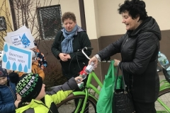 Chłopiec w kolorowej kurtce podaje butelkę wody mineralnej uśmiechniętej kobiecie. Stoi ona obok zielonego roweru. W ręce trzyma torebkę i torbę na zakupy. Na tle budynku widać nauczycielkę i z lewej strony stojące przedszkolaki.
