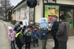 Przed sklepem z napisem: Delikatesy Centrum stoi grupa przedszkolaków wraz z nauczycielką. Przed nimi, zwrócony do nich twarzą stoi mężczyzna w ciemnej kurtce   i czarnej czapce. Dzieci trzymają transparenty.
