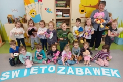 Sala przedszkolna . Grupa dzieci ubrana na kolorowo stoi  obok siebie przed zestawem szafek przedszkolnych . W grupie stojącej widać osobę dorosłą – wychowawczynię z kolorową szmacianą lalką. Przed grupą stojącą 8 dzieci siedzi na podlodze i trzyma na kolanach swoją zabawkę. Przed dziećmi napis z białych liter na niebieskim tle- ŚWIĘTO ZABAWEK.