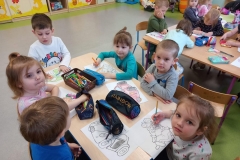 Przy stoliku grupa dzieci  . Dzieci kolorują obrazki konturowe zabawek . Na stolikach rozłożone kredki i piórniki z przyborami do kolorowania.