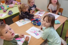 Przy stoliku grupa dzieci  . Dzieci kolorują obrazki konturowe zabawek . Na stolikach rozłożone kredki i piórniki z przyborami do kolorowania.