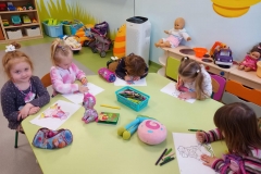 Przy stoliku grupa dzieci  . Dzieci kolorują obrazki konturowe zabawek . Na stolikach rozłożone kredki i piórniki z przyborami do kolorowania.