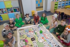 Dzieci siedzą w sali przedszkolnej. Przed nimi leży duża plansza- gra "Wyprawa do lasu". Na planszy ilustracje i polecenia dotyczące zasad gry.