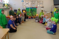 Na sztaludze zawieszony obraz przedstawiający las i jego mieszkańców. Z obydwu stron na podłodze siedzą dzieci z opaskami zwierząt na głowie. Za nimi stoi 4 chłopców i 1 dziewczynka w zielonych pelerynach.