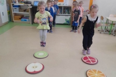 Dwie dziewczynki skaczą jak piłeczki po okrągłych matach. W tyle dzieci stoją w dwóch rzędach.