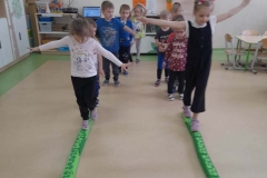 Dwie dziewczynki idą po zielonym torze, ręce trzymają w bok, ćwiczą równowagę. Reszta dzieci stoi  za nimi w dwóch rzędach.