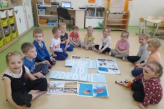 Dzieci siedzą w półkole na podłodze, na podłodze widać napis sport to zdrowie, wokół napisu kolorowe dyscypliny sportu.