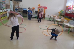 Na podłodze rozłożone obręcze, dzieci skaczą do nich, do ostatniej wchodzą i przekładają przez siebie, w tyle dwa rzędy dzieci.