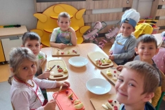 Grupa dzieci ubrana na kolorowo; w fartuszkach . Jeden z chłopców na czepek na głowie. – 5 chłopców i jedna dziewczynka siedzią przy prostokatnym stoliku. Przed każdym dzieckiem leży deska do krojenia , a na niej owoce . Dzieci kroją na deskach owoce. Za dziećmi półka na zabawki w kształcie słońca. Za półką obudowa kaloryfera i część okien , a na nich wiosenne dekoracje.